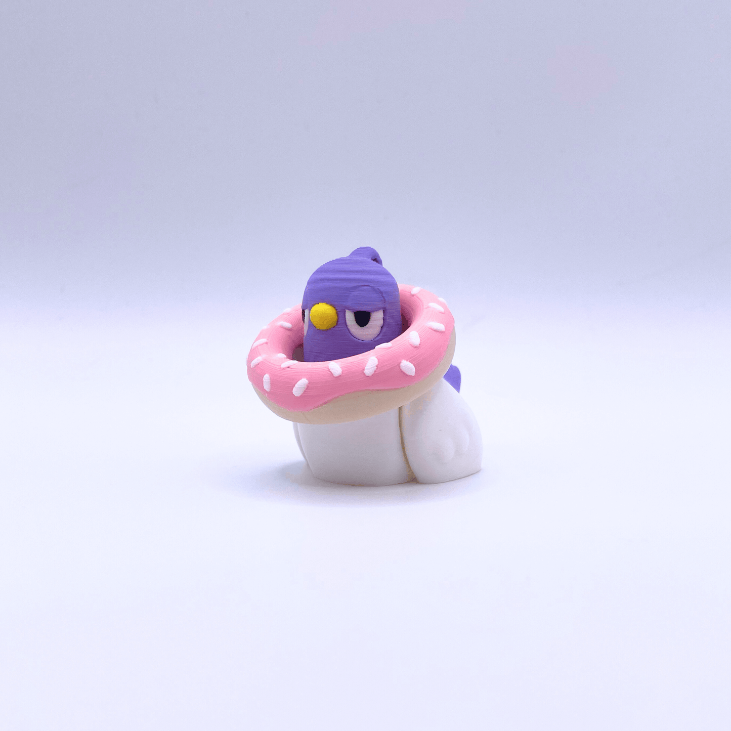 Small pigeon with a purple head and white body, wearing a pink donut with white sprinkles on a light purple background.