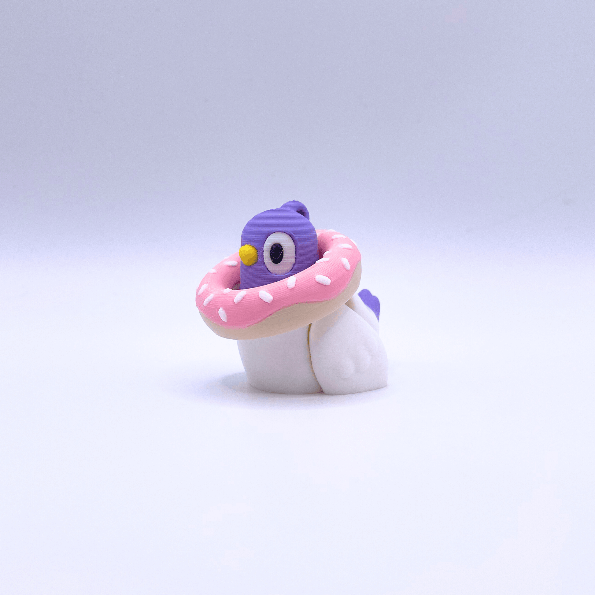 Toy figure of a pigeon with a purple head and pink donut-like body on a light gray background