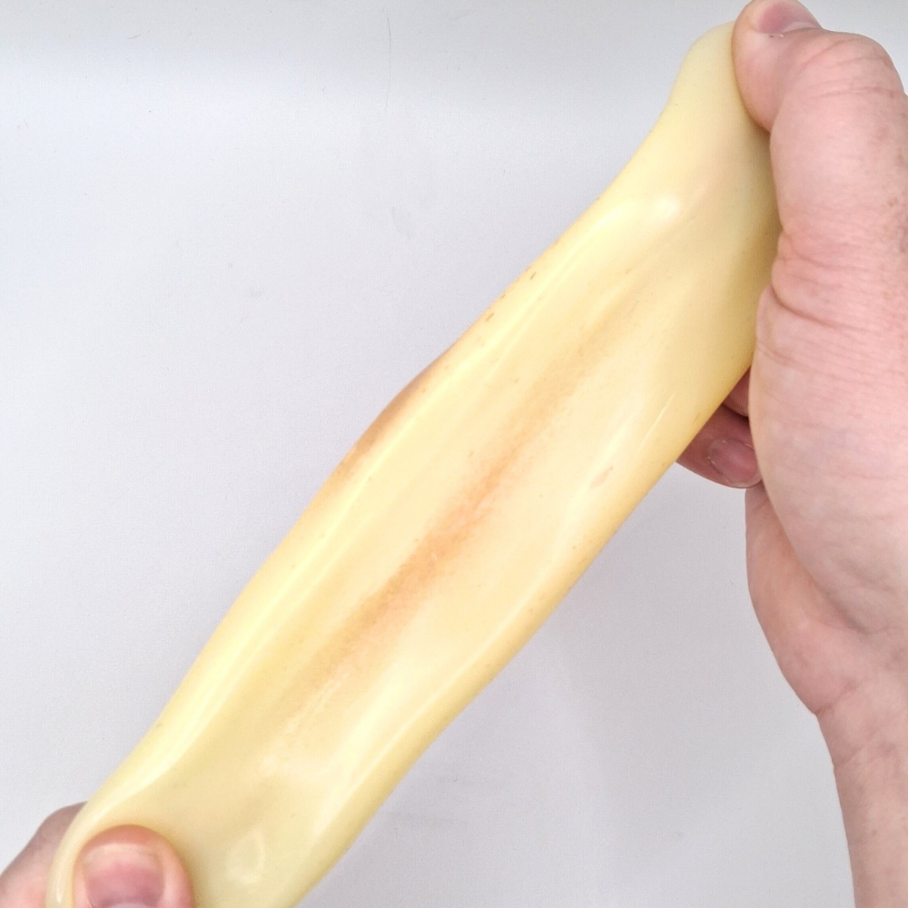 A hand holding a long, pale yellow, potato squish against a plain light grey background.